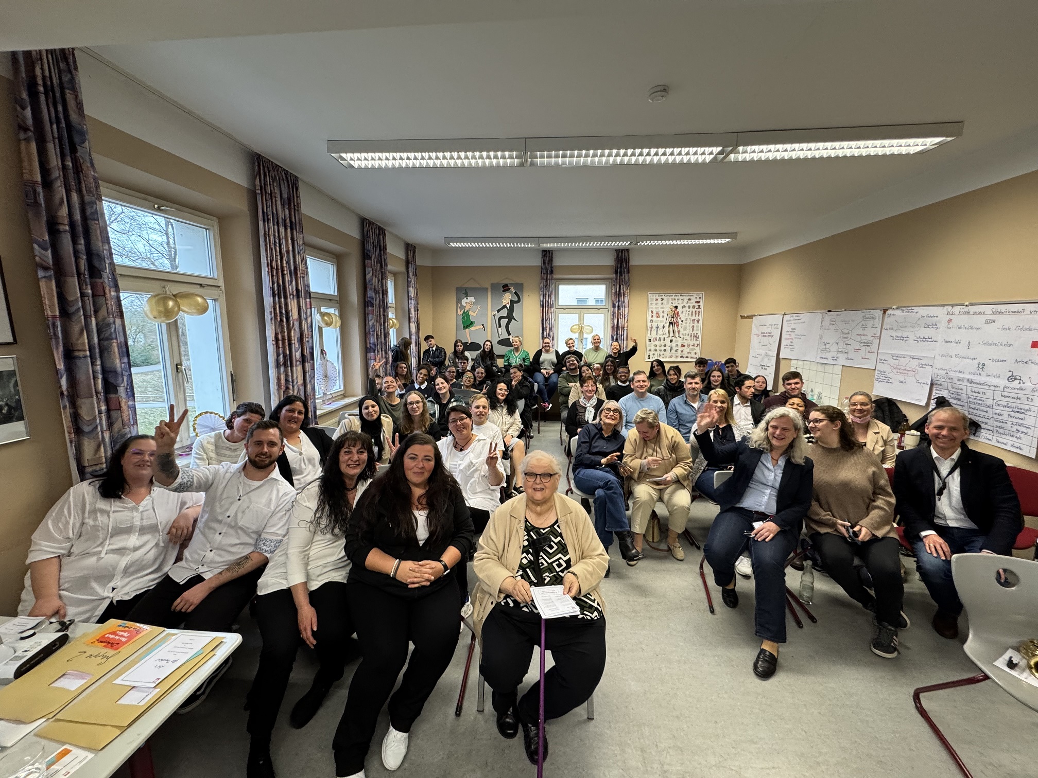 Eine große Gruppe von Menschen unterschiedlichen Alters und Geschlechts sitzt dicht beieinander in einem hell erleuchteten Klassenzimmer. Viele lächeln oder winken in die Kamera. An den Wänden hinter ihnen sind Plakate und Schriftzüge zu sehen.