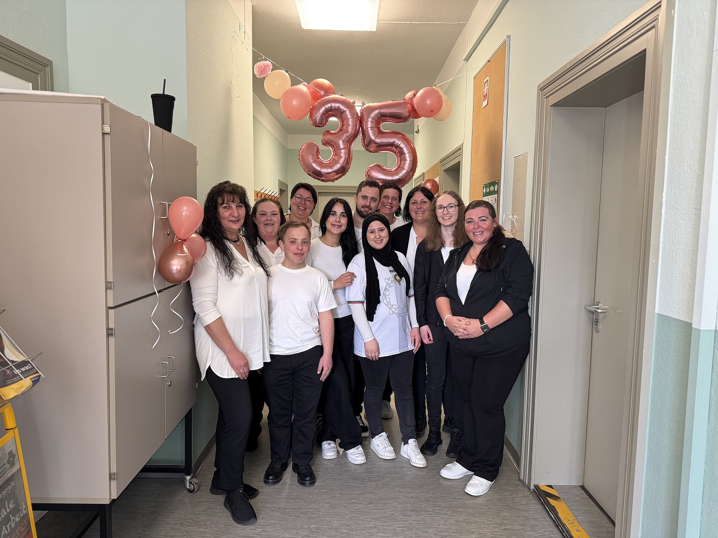 Eine Gruppe von zehn Personen, überwiegend in Schwarz und Weiß gekleidet, steht lächelnd in einem Flur unter der rosa Ballonnummer „35“. Der Flur ist mit Ballons dekoriert und die Gruppe scheint ein Ereignis zu feiern.