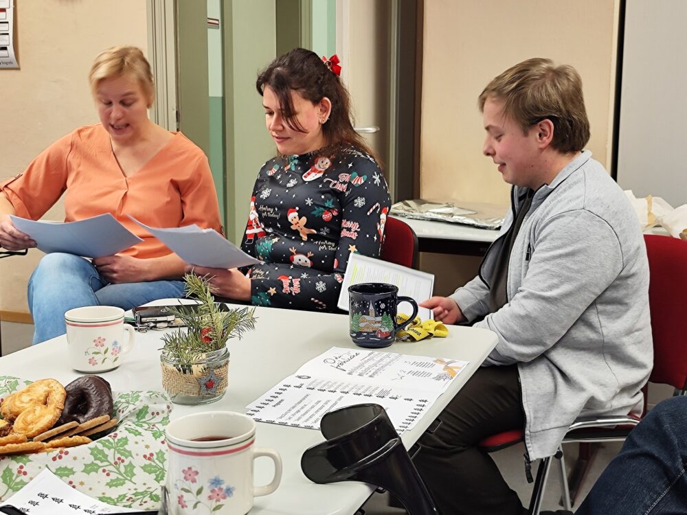 Drei Personen sitzen zusammen an einem Tisch, lesen Zeitung und lächeln. Auf dem Tisch stehen Kaffeetassen, Gebäck und eine kleine Topfpflanze. Die Szene suggeriert ein zwangloses Treffen oder Zusammensein in gemütlicher Atmosphäre.