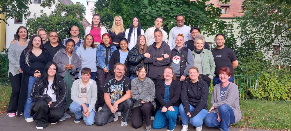 Eine Gruppe von 28 Personen unterschiedlichen Alters, Geschlechts und ethnischer Zugehörigkeit posiert im Freien vor grünen Bäumen und Gebäuden. Die meisten lächeln, einige stehen, einige knien oder sitzen vorne.