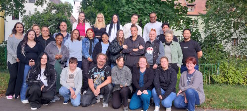 Eine Gruppe von 28 Personen unterschiedlichen Alters, Geschlechts und ethnischer Zugehörigkeit posiert im Freien vor grünen Bäumen und Gebäuden. Die meisten lächeln, einige stehen, einige knien oder sitzen vorne.