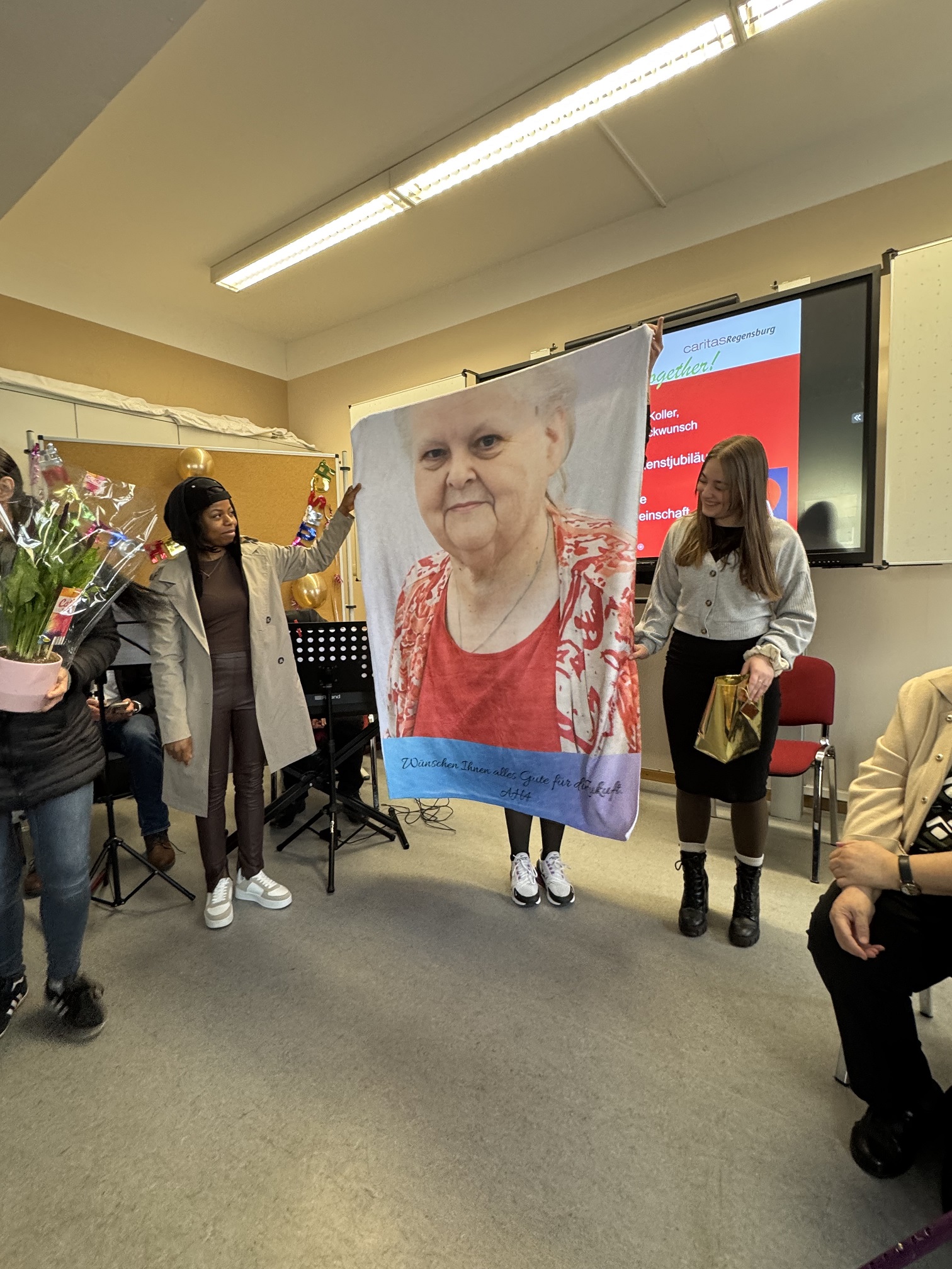 Drei Frauen stehen in einem Raum und halten eine große Decke mit dem Porträt einer älteren Frau in den Händen. Die Gruppe ist von Menschen, Blumen und Musikinstrumenten umgeben, was an eine Feier oder eine Gedenkveranstaltung erinnert.