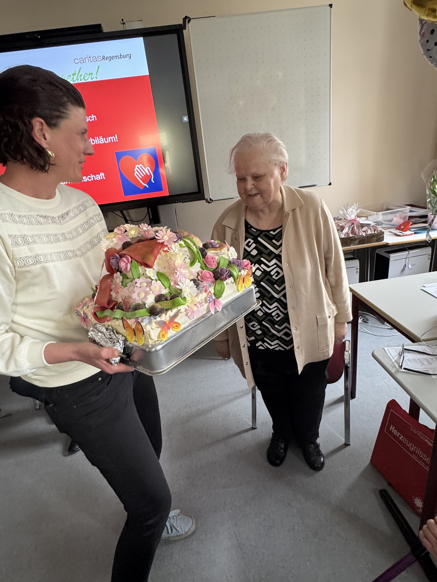 Eine Frau überreicht einer lächelnden älteren Frau einen großen, bunten Kuchen, der mit Blumen und Erdbeeren dekoriert ist. Sie befinden sich in einem Raum mit einer weißen Tafel, einem Bildschirm und einem Tisch mit Geschenken im Hintergrund.