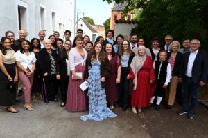 Eine Gruppe von etwa 30 Personen unterschiedlichen Alters posiert im Freien, viele von ihnen in formeller oder traditioneller Kleidung, und lächelt gemeinsam unter Bäumen neben weißen Gebäuden.