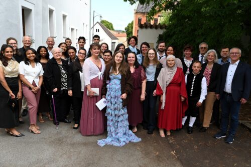 Eine Gruppe von etwa 30 Personen unterschiedlichen Alters posiert im Freien, viele von ihnen in formeller oder traditioneller Kleidung, und lächelt gemeinsam unter Bäumen neben weißen Gebäuden.