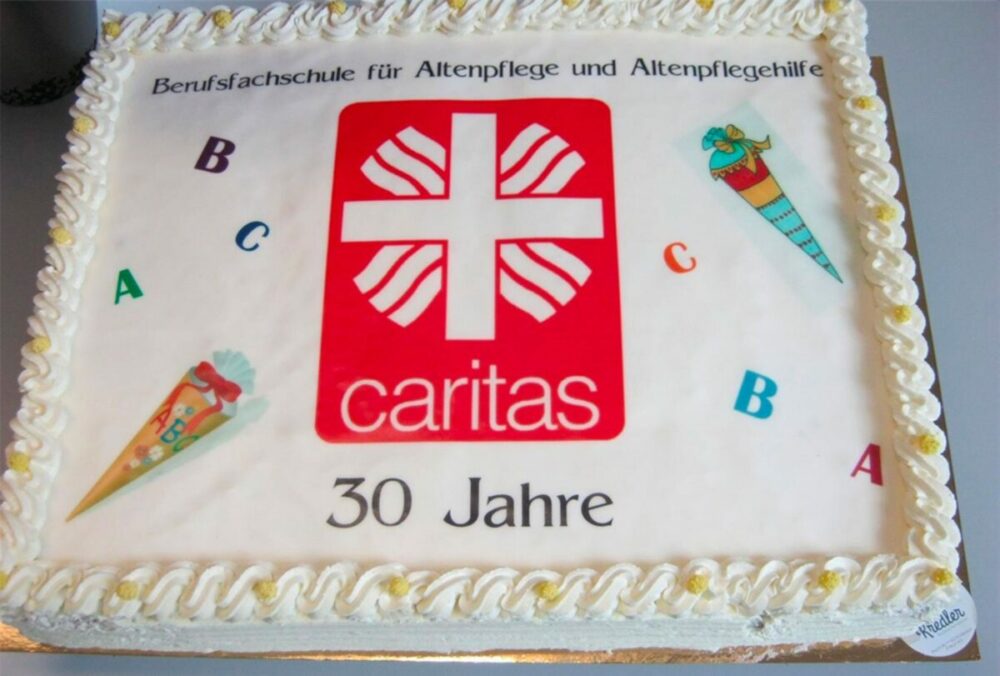 Eine rechteckige Torte, verziert mit dem Caritas-Logo, dem deutschen Text „Berufsfachschule für Altenpflege und Altenpflegehilfe“, 30 Jahre, sowie farbenfrohen Illustrationen und Buchstaben zum Thema Schule auf weißem Zuckerguss mit paspeliertem Rand.