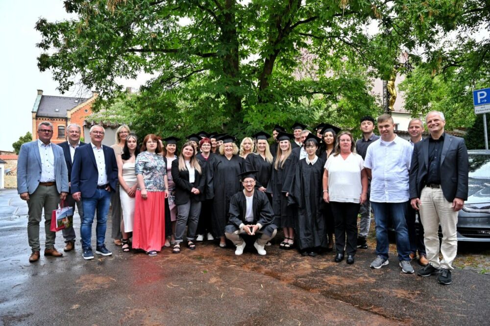 Eine Gruppe von Menschen, darunter Absolventen in Talar und Barett, steht draußen auf einem nassen Bürgersteig unter einem großen Baum und lächelt in die Kamera. Einige sind formell gekleidet, andere leger.