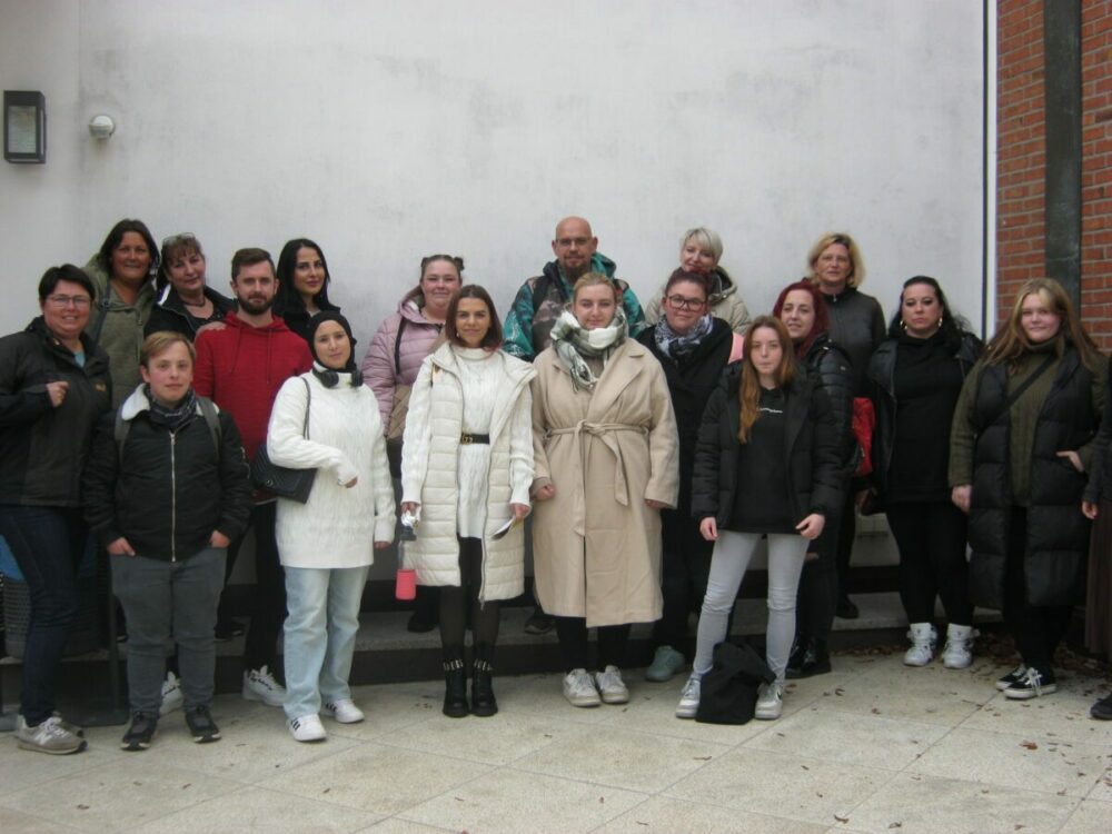 Eine Gruppe von neunzehn Personen, Erwachsene und Kinder, steht zusammen draußen vor einer weißen Wand und einer gemauerten Ecke, in Winterkleidung wie Mänteln und Schals gekleidet, und posiert für ein Gruppenfoto.