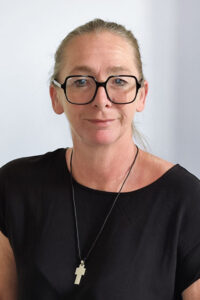 Eine Frau mit hellbraunem, zurückgebundenem Haar, einer schwarz umrandeten Brille, einem schwarzen Oberteil und einer Halskette mit einem Kreuzanhänger steht vor einem schlichten, hellen Hintergrund.