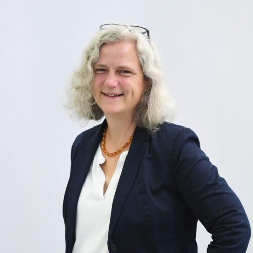 Eine Frau mit welligem grauem Haar, einer Brille auf dem Kopf, einem marineblauen Blazer, einer weißen Bluse und einer Bernsteinkette lächelt vor einem schlichten hellen Hintergrund in die Kamera.