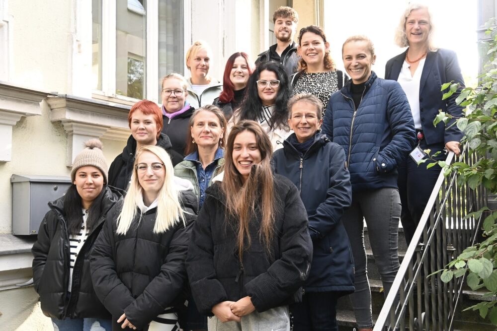 Eine Gruppe von vierzehn Personen, überwiegend Frauen, steht lächelnd auf den Stufen einer Außentreppe neben einem Gebäude. Die meisten tragen Jacken, manche haben Beanies auf. Die Stimmung ist freundlich und locker.